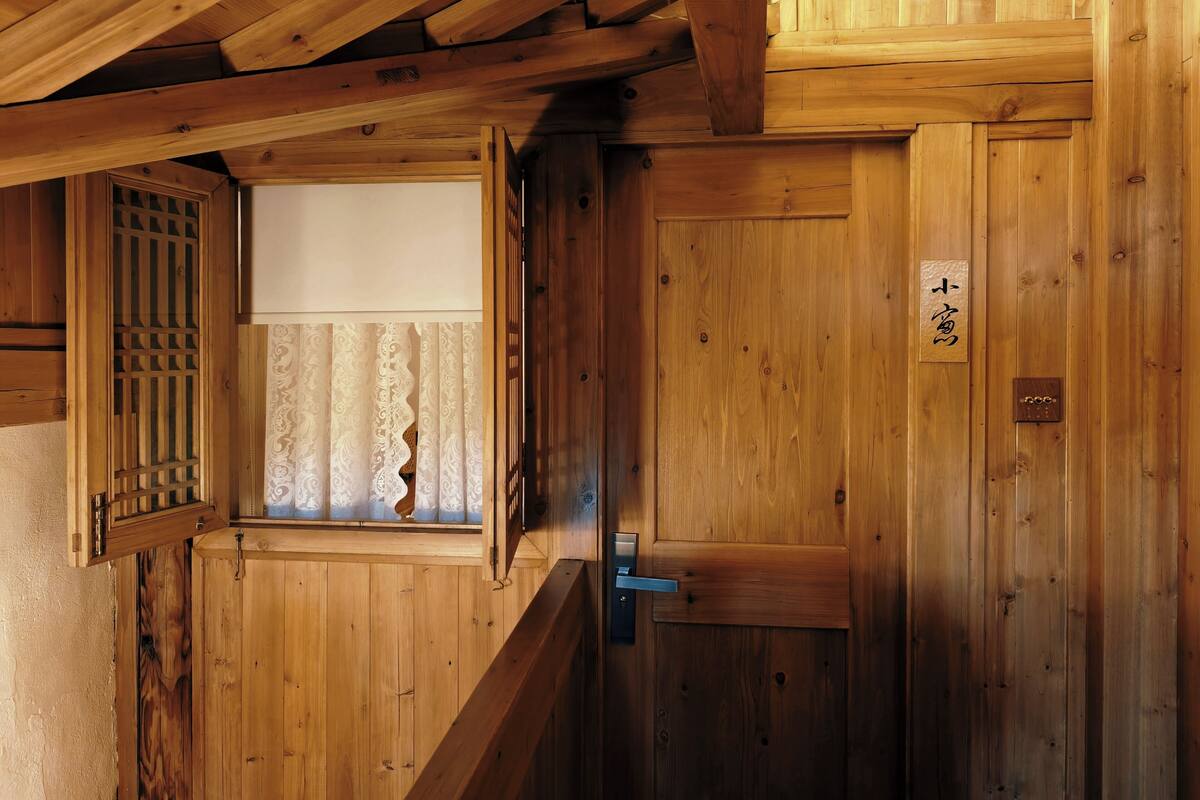 Un interior de madera con un puertas corredera, una ventana con cortinas estampadas y un letrero de madera con caracteres japoneses.