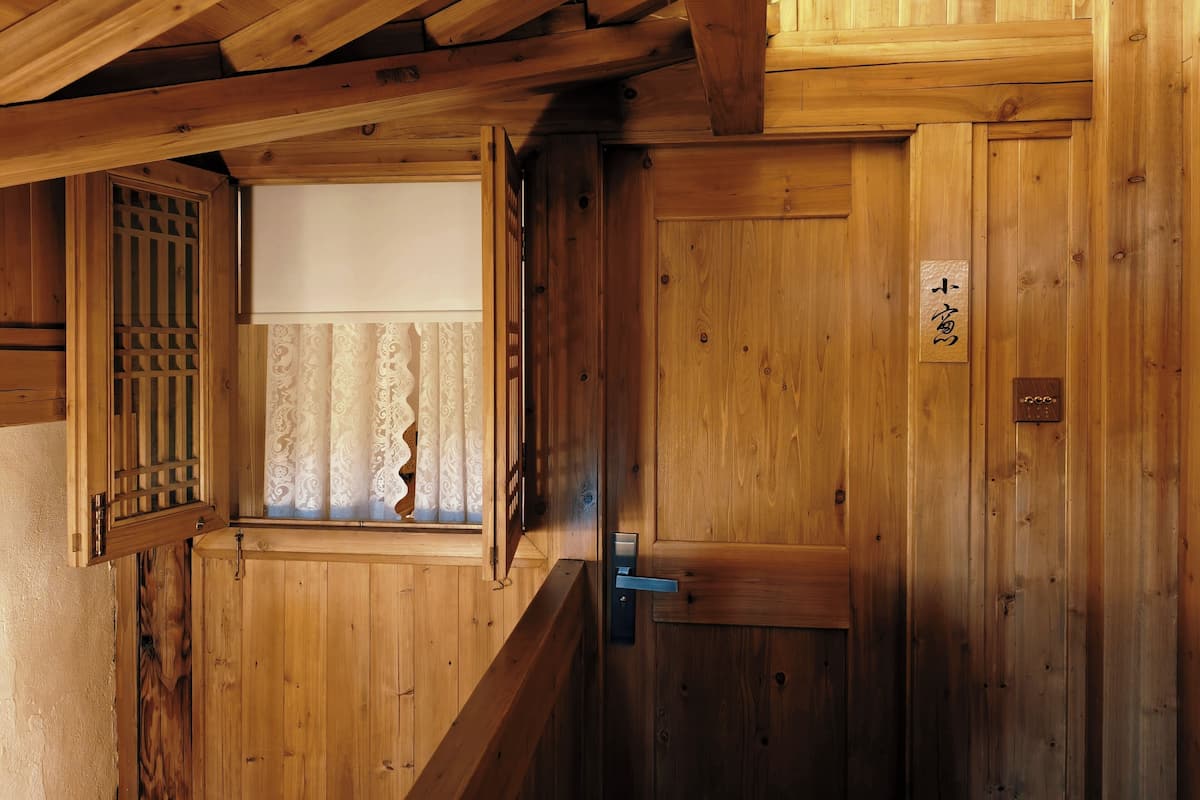 A wooden interior with a sliding door, a window with patterned curtains, and a wooden sign with Japanese characters.