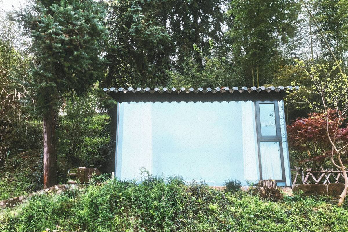 Una pequeña estructura azul claro con techo de tejas, rodeada de abundante vegetación y árboles.