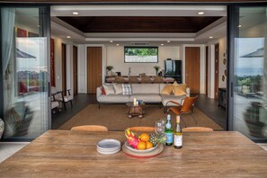 A modern living room with a dining area, a sofa, a coffee table, and a TV. - VILLA AZURE - 4BR LUXURY VILLA (ตำบลเกาะพะงัน)