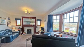 A spacious living room with a fireplace, leather sofas, a coffee table, and large windows offering a view. - Plover Apartment - UK51129 (Carradale)