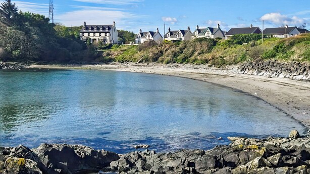 A coastal area with a rocky shoreline, a calm bay, and a row of houses with a prominent white building on the left. - Plover Apartment - UK51129 (Carradale)