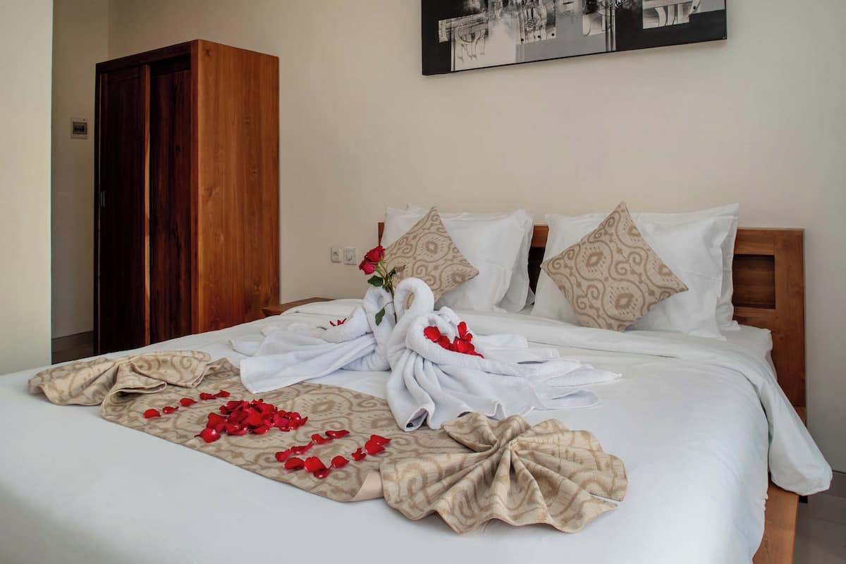 Una cama bien hecha con una toalla y pétalos de rosa, un armario de madera y un cuadro en la pared.