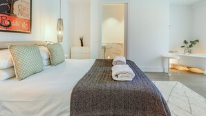 Una habitación de hotel moderna con una cama grande, una mesita de noche con una planta, un baño con espejo y una obra de arte colgada en la pared.