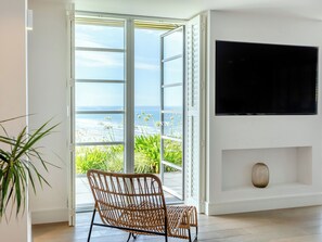 Ein Zimmer mit an der Wand befestigtem Fernseher, einem Korbstuhl und einer Glastür zum Balkon mit Meerblick.
