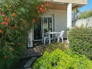 A patio area with a table and chairs, a glass door leading to a room, and a small garden with greenery and a tree with red flowers. - Relaxing Corsica Stay With Terrace (Linguizzetta)