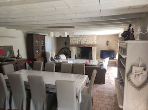 A dining area with a table set for four, surrounded by chairs. There is a stone fireplace with a mantel, a television on a stand, and a cabinet with glass doors. - Very large farmhouse in a quiet hamlet near Auray and Carnac. (Ploemel)
