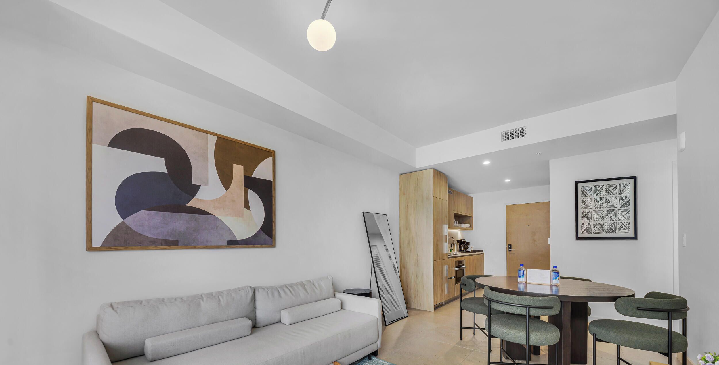 Ein modernes Wohnzimmer mit einem grauen Sofa, einem Esstisch mit Stühlen, einer Küchenzeile und abstrakter Kunst an den Wänden.