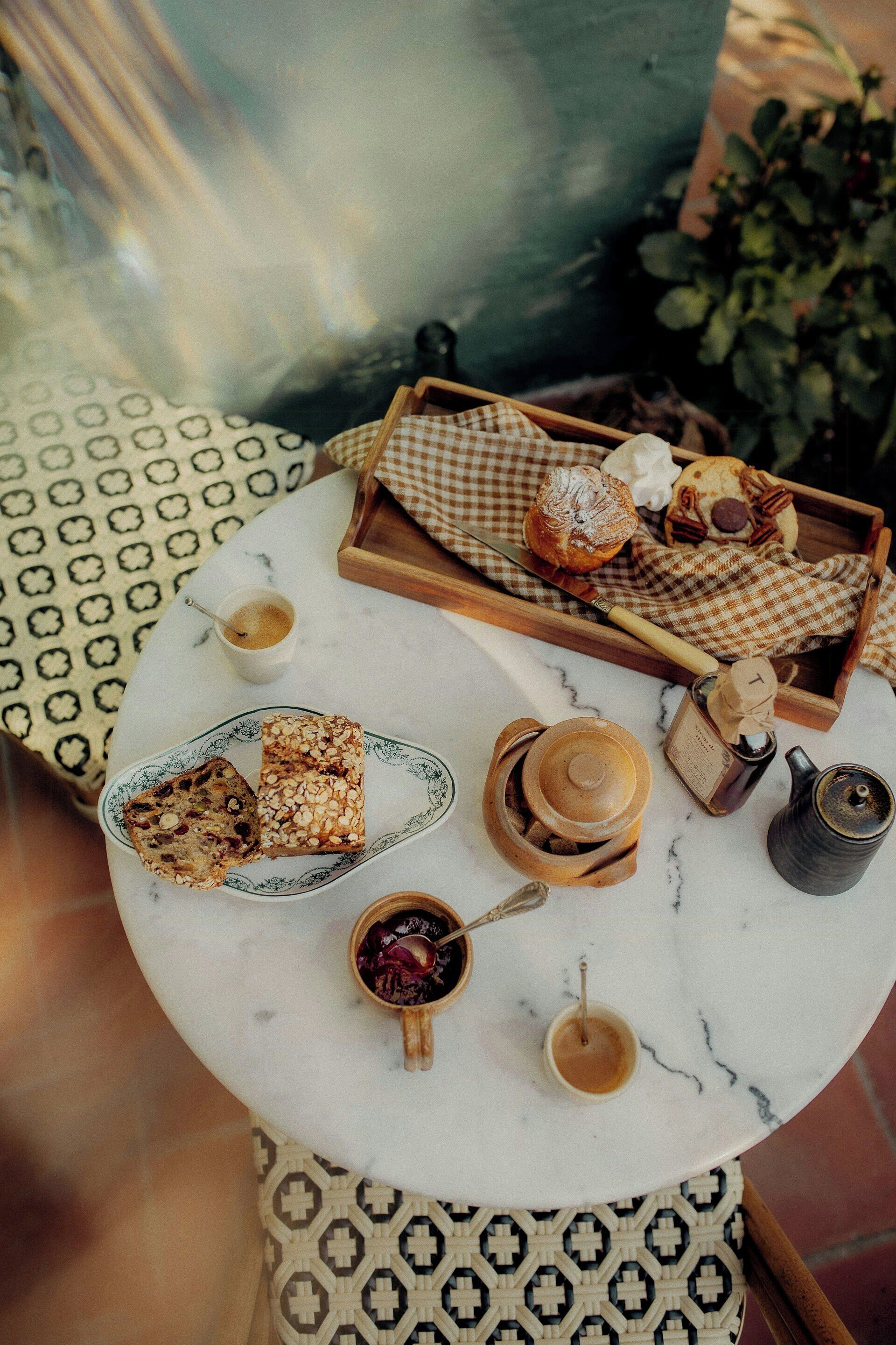 A round table with a checkered cloth, a tray with pastries, a cup of coffee, and a small bottle of jam.