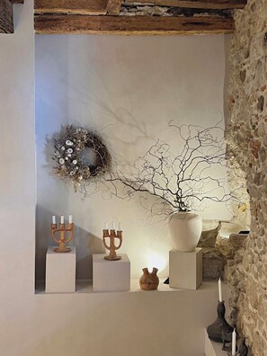 Une piĂšce avec un mur en pierre, un plafond en bois, une Ă©tagĂšre blanche ornĂ©e dâun chandelier, dâun vase et dâune couronne.