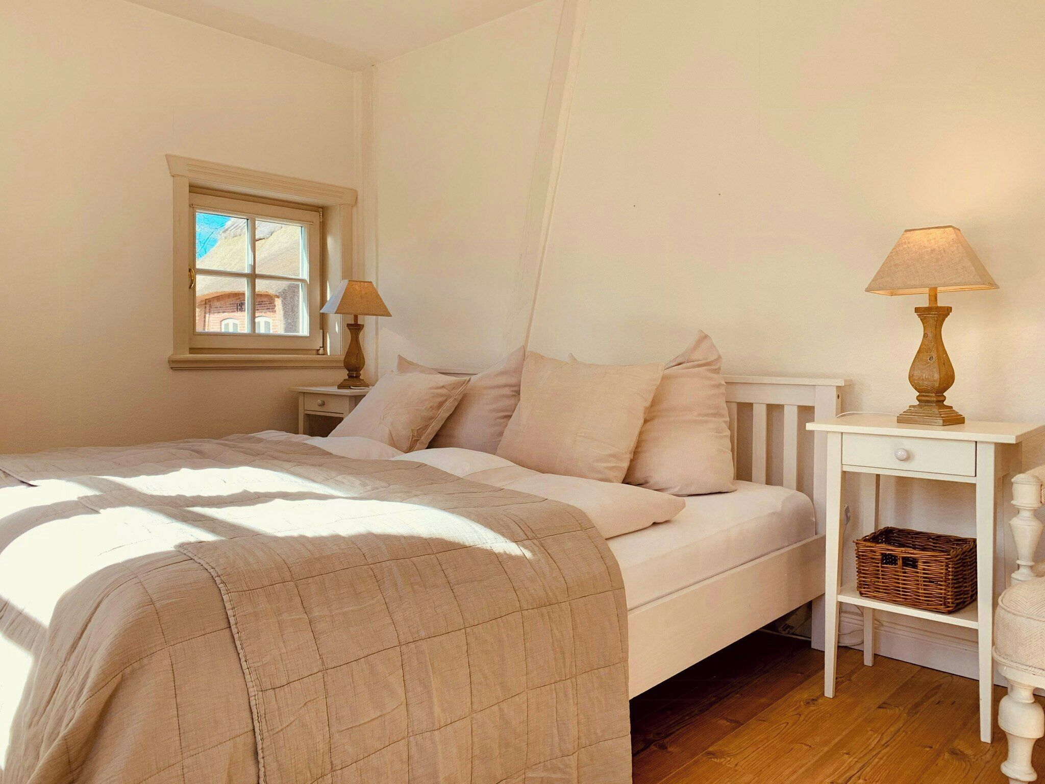 Ein Schlafzimmer mit einem Bett, einem Nachttisch mit Lampe, einem Fenster und einem Beistelltisch mit Korb.