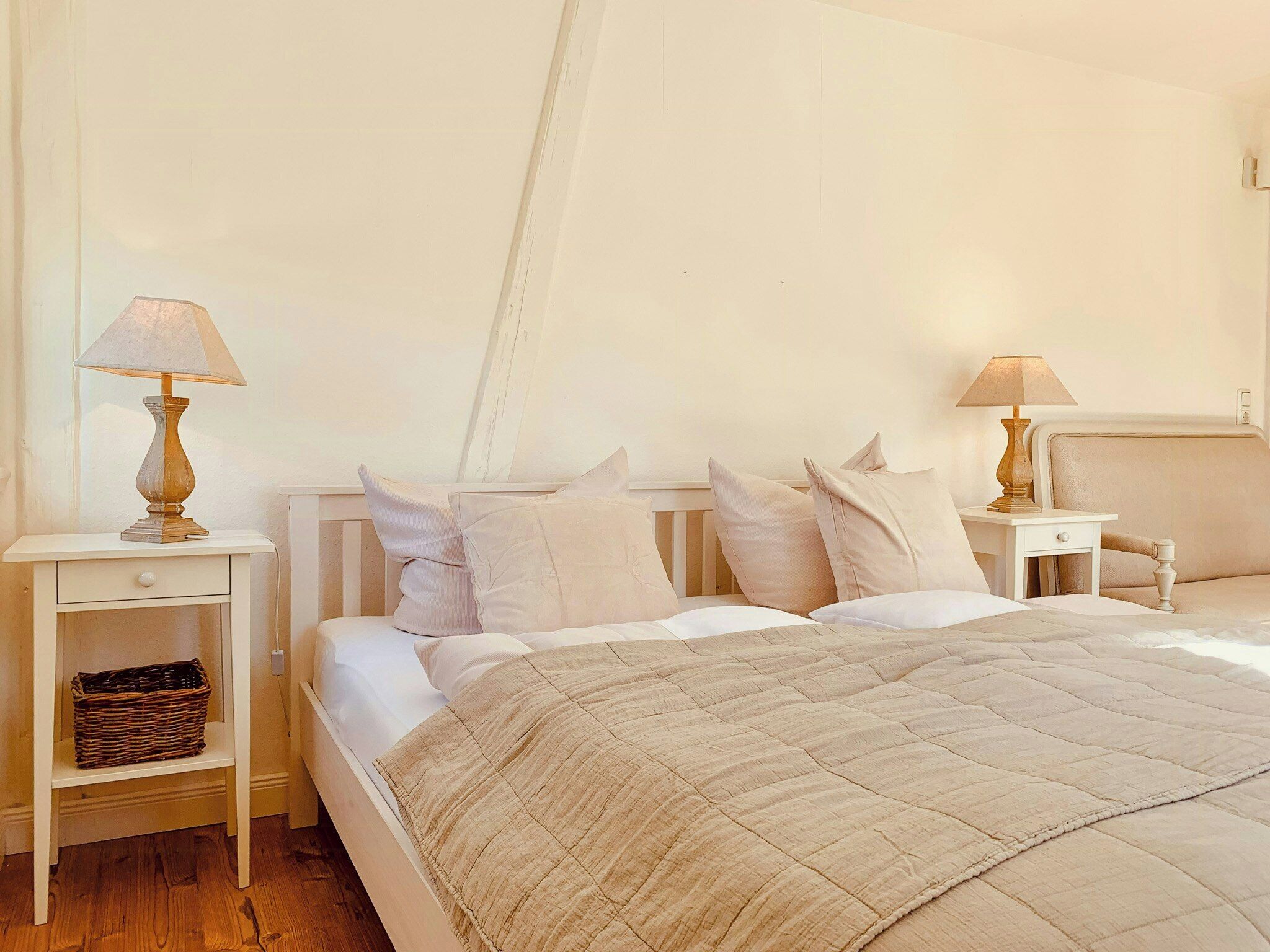 A bedroom with a bed, two bedside tables, a lamp on each table, a nightstand with a basket, and a headboard.