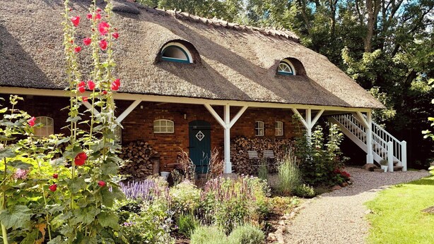 Et bindingsværkshus med stråtækt tag, en overdækket veranda, en trappe og en have med forskellige planter.
