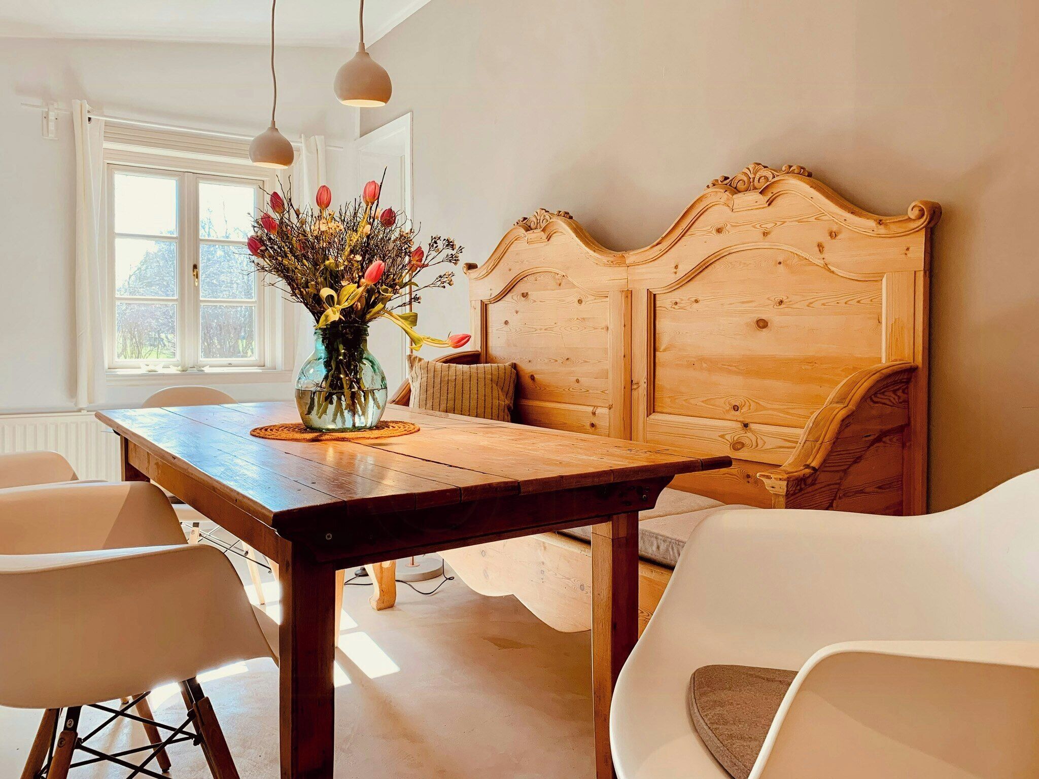 A dining area with a wooden table, white chairs, a vase with flowers, and a wooden headboard.