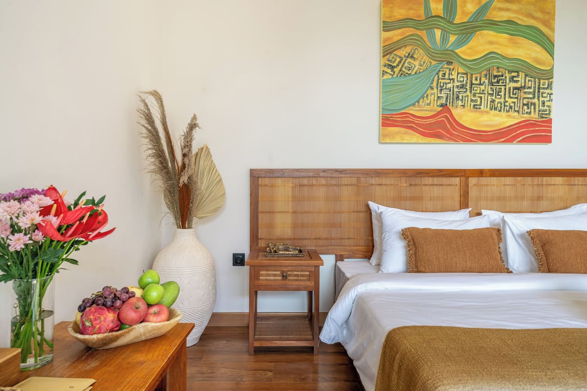 Ein Hotelzimmer mit einem Bett, einem Nachttisch, einer Vase mit Blumen, einer Obstschale und einem Wandbild.