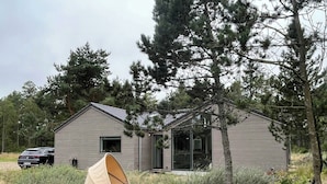 Une petite cabane d’un seul étage, dotée d’un espace extérieur abrité avec des sièges, et entourée d’arbres et de végétation.