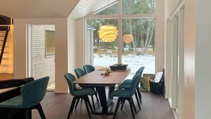 Un espace repas avec une table et des chaises en bois, de grandes fenêtres et une vue sur l’extérieur.