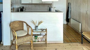 A kitchen with wooden cabinets, a white island, a hanging light fixture, a wicker chair, and a small table with a vase. - Bright holiday home close to sea near Blokhus (Blokhus)