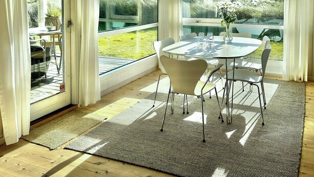 A room with a wooden ceiling, large windows with curtains, a round table with chairs, and a view of greenery outside. - Bright holiday home close to sea near Blokhus (Blokhus)