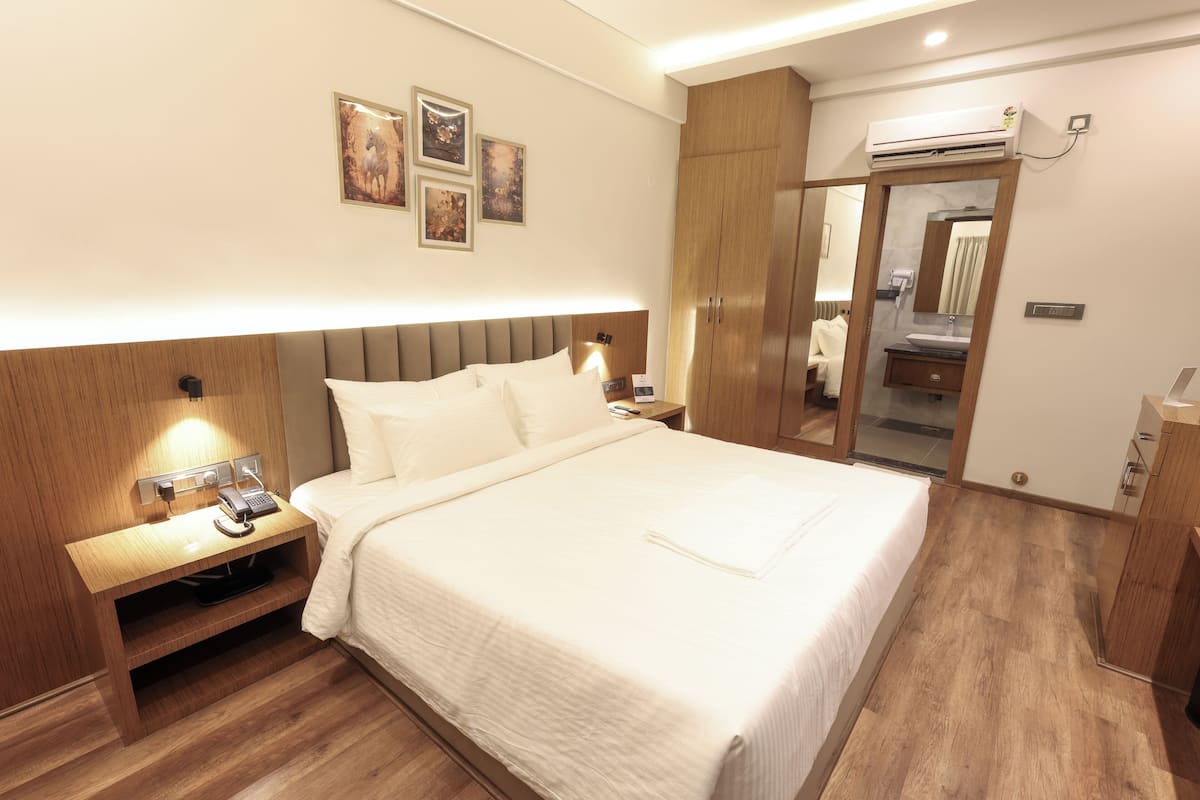 Quarto de hotel com uma cama grande, cabeceira de madeira, mesas de cabeceira, um criado-mudo, um espelho, um guarda-roupa e um aparelho de ar condicionado.