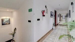 Un pasillo con una señal de salida de incendios, un extintor, cuadros en las paredes y plantas en macetas.