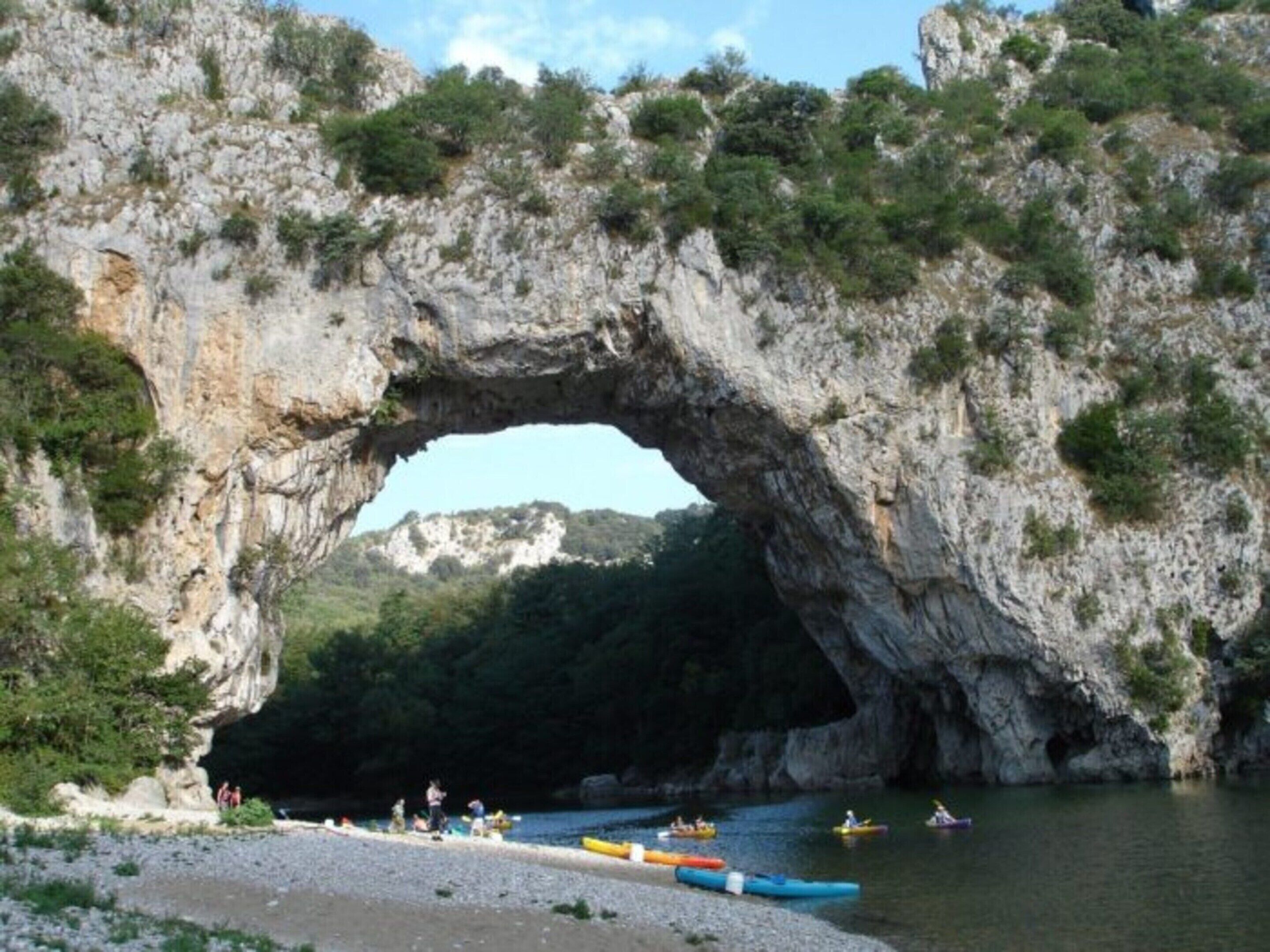 A natural rock arch formation with a river flowing through it, surrounded by greenery and a clear sky.