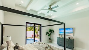 Quarto com cama, televisão, cômoda, ventilador de teto e vista para uma piscina.