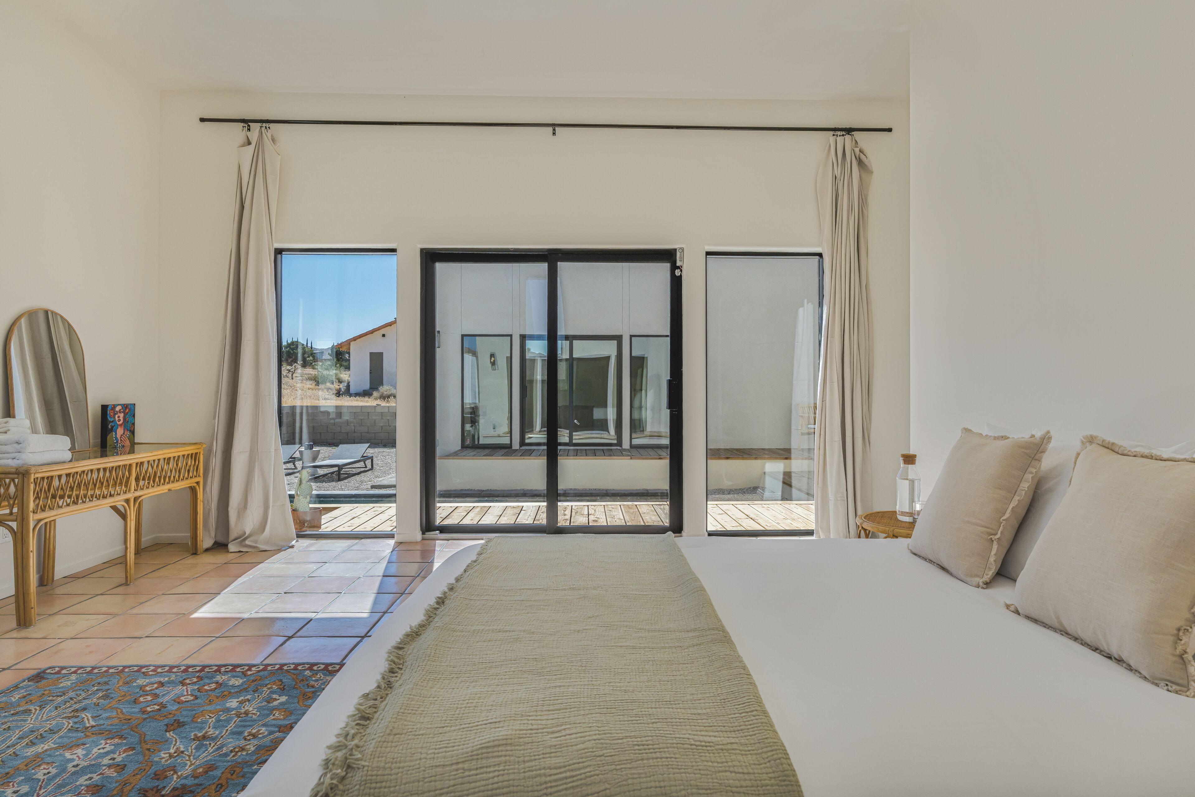 Un dormitorio con cama, vista a través de una puerta corrediza de vidrio, una mesita de noche con lámpara y una ventana con cortinas.