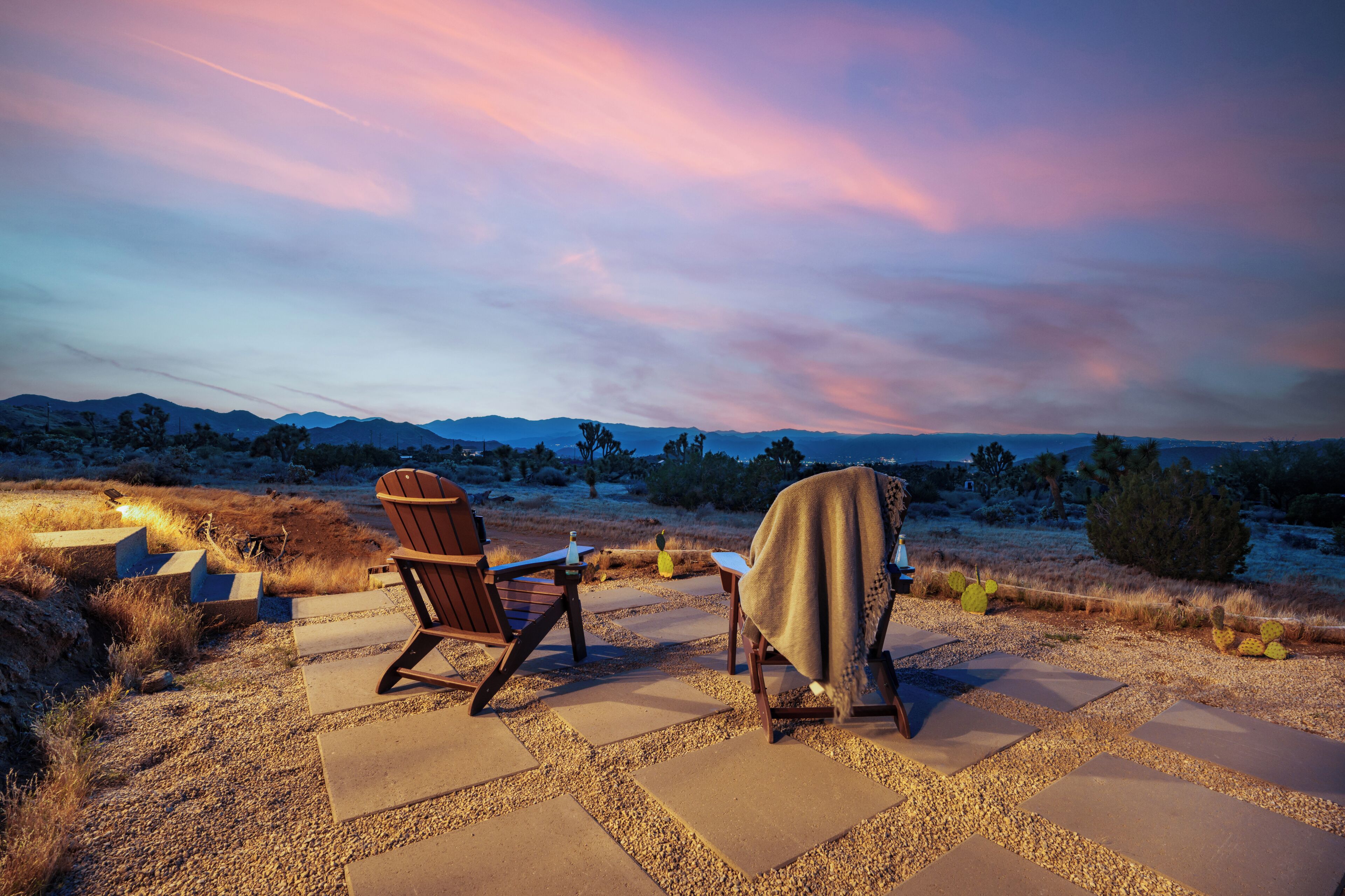 A patio with two Adirondack chairs, a blanket, and a view of the desert landscape.