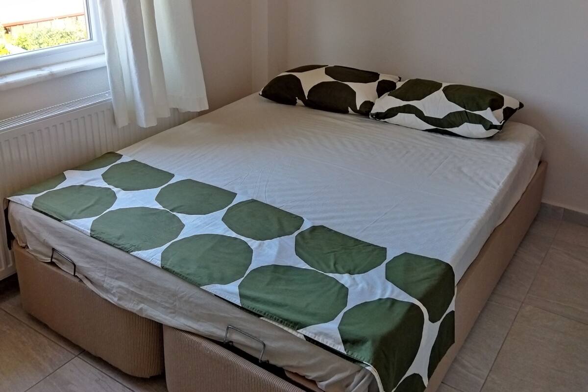 A bed with a green and white patterned bedspread, a pillow with a similar pattern, and a storage ottoman.