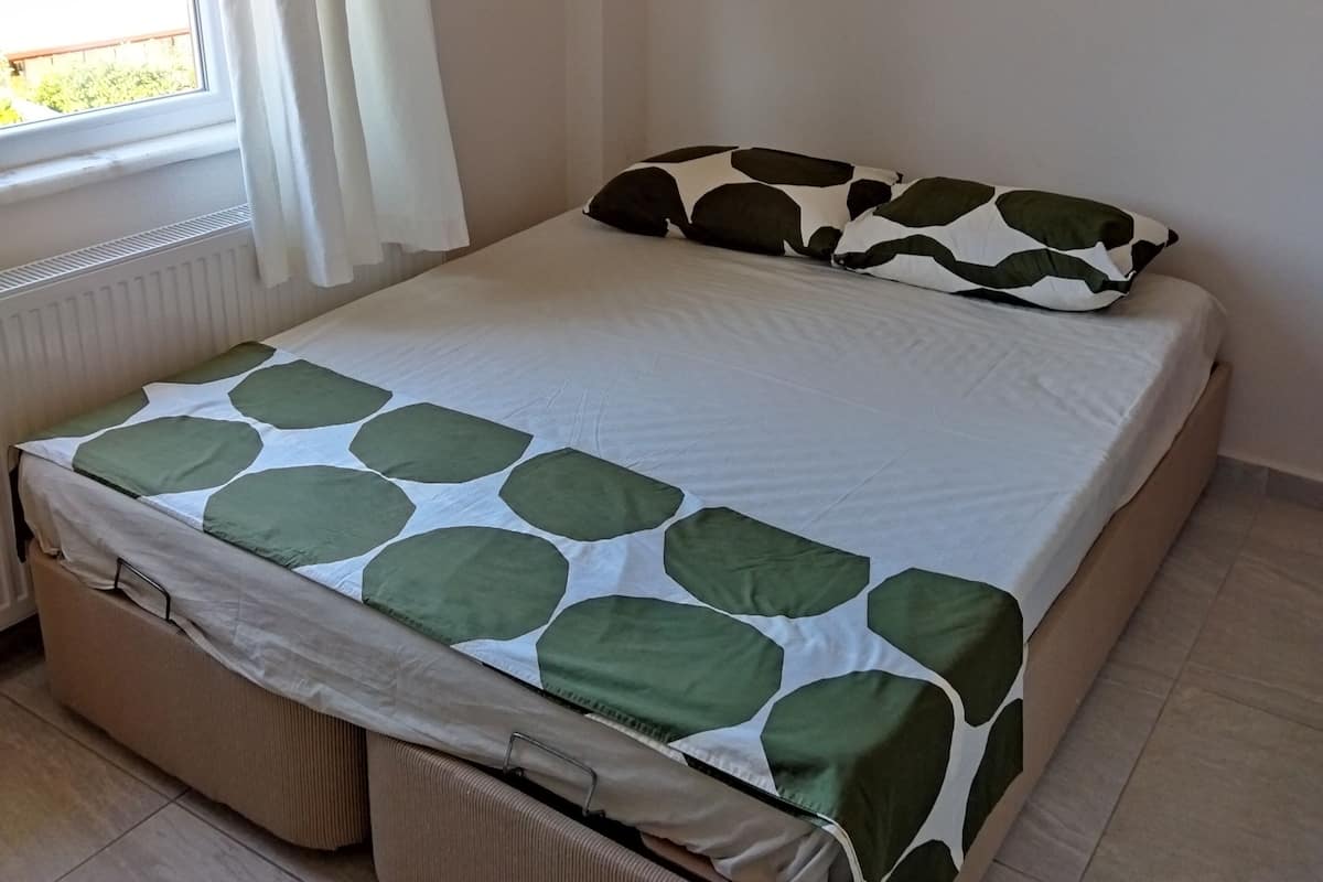 A bed with a green and white patterned bedspread, a pillow with a similar pattern, and a storage ottoman.