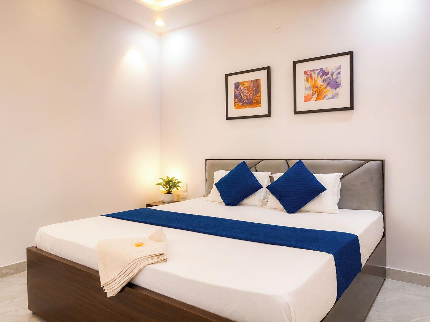 A hotel room with a bed, a bedside table with a lamp, and two framed artworks on the wall.