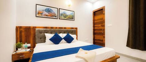 A hotel room with a large bed, wooden headboard, two wall-mounted pictures, a bedside table with a bottle and glasses, and a dark curtain.