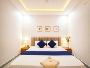 A hotel room with a large bed, two bedside tables, a ceiling light, and three framed pictures on the wall. - Maa Durga Dham (Vrindavan)