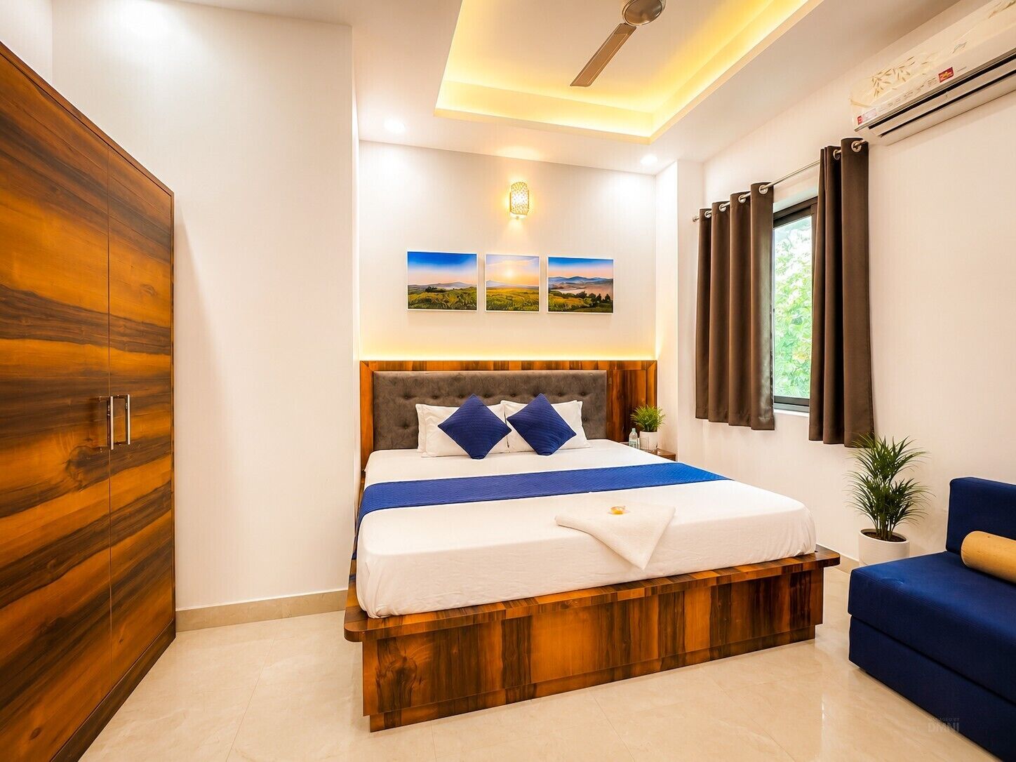 A bedroom with a large wardrobe, a bed with a wooden headboard, a blue sofa, and a window with curtains.