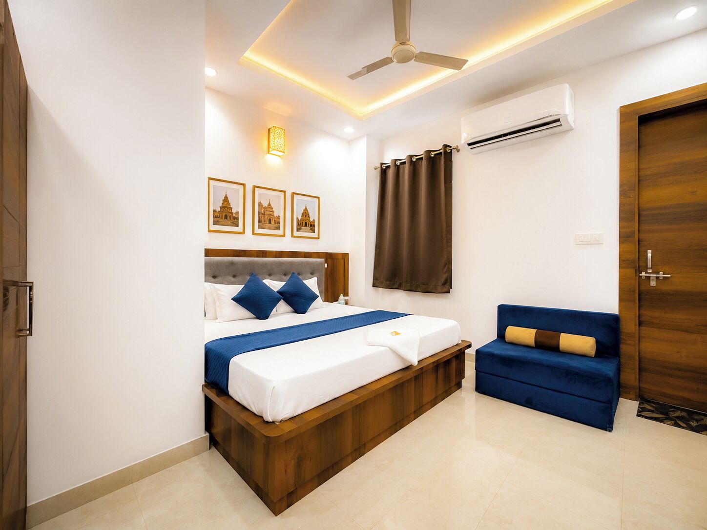 A hotel room with a bed, a blue sofa, a ceiling fan, an air conditioning unit, and framed pictures on the wall.