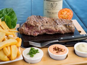 Ein Steak mit Pommes, eine Flasche BOURGOMY und zwei kleine Schälchen Soße auf einem Holzbrett.