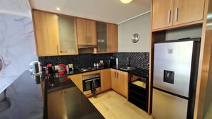 A modern kitchen with wooden cabinets, a stainless steel refrigerator, a black countertop, and a tiled backsplash. - Waterfront Deco apartment 2 sleeper lake views (Cape Town)