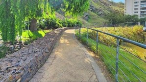 Un chemin bordé d’un côté par un mur de pierres et de l’autre par une rambarde métallique, qui mène à une zone herbeuse plantée d’arbres.