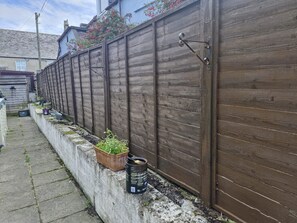 Una cerca de madera con un portón metálico, una jardinera de concreto con plantas y un bote negro en el suelo.