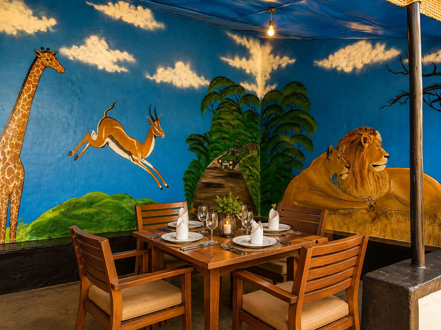 A dining area with a mural of a giraffe, antelope, and lion, wooden chairs, and a table set for two with plates, glasses, and cutlery.