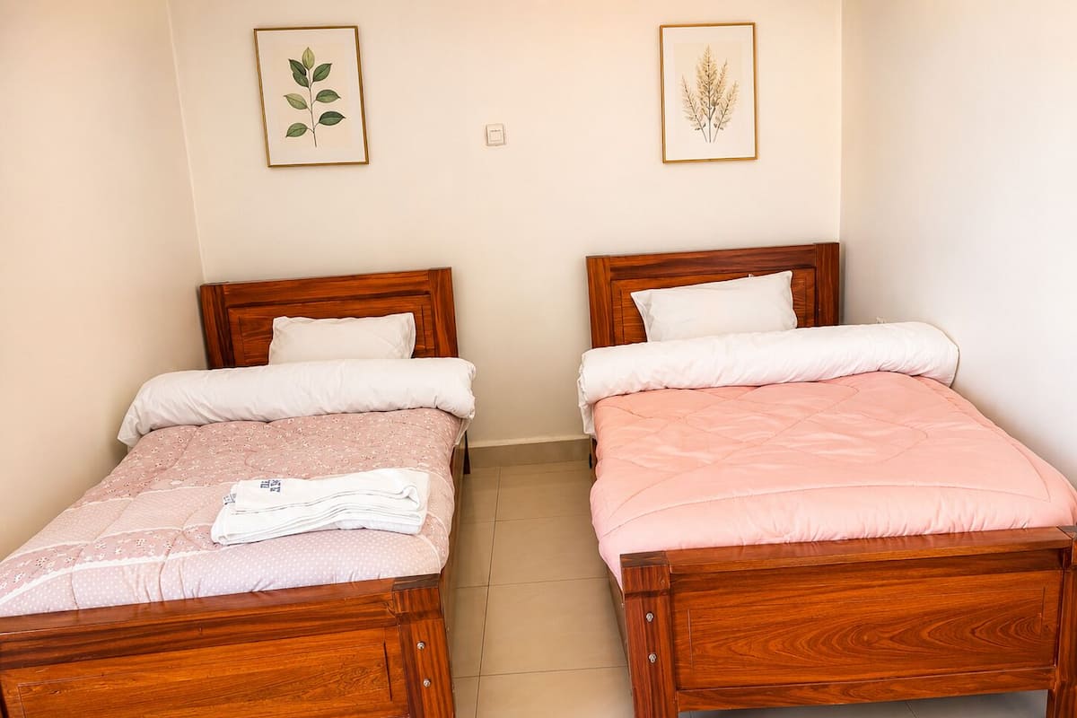 Dos camas de madera con ropa de cama blanca y rosa, una toalla pequeña e impresiones botánicas enmarcadas en la pared.