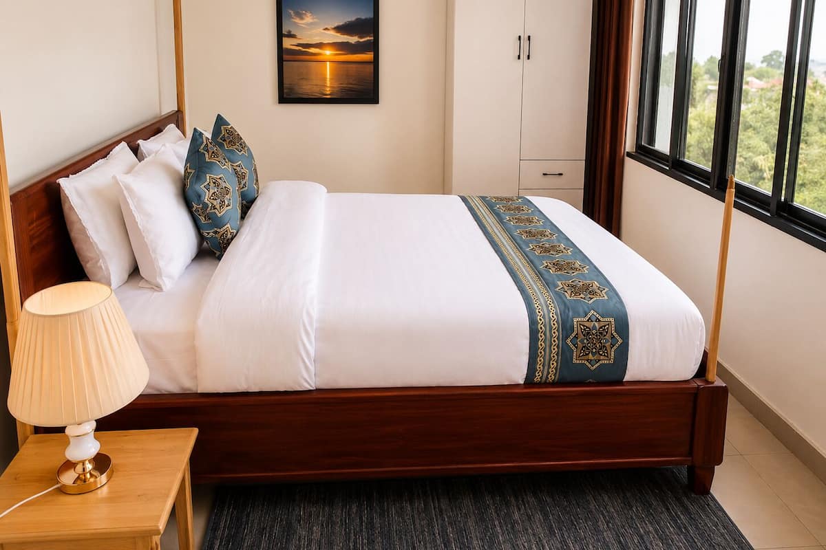 Una cama bien hecha con una colcha estampada, una lámpara de noche y vistas a un atardecer a través de la ventana.
