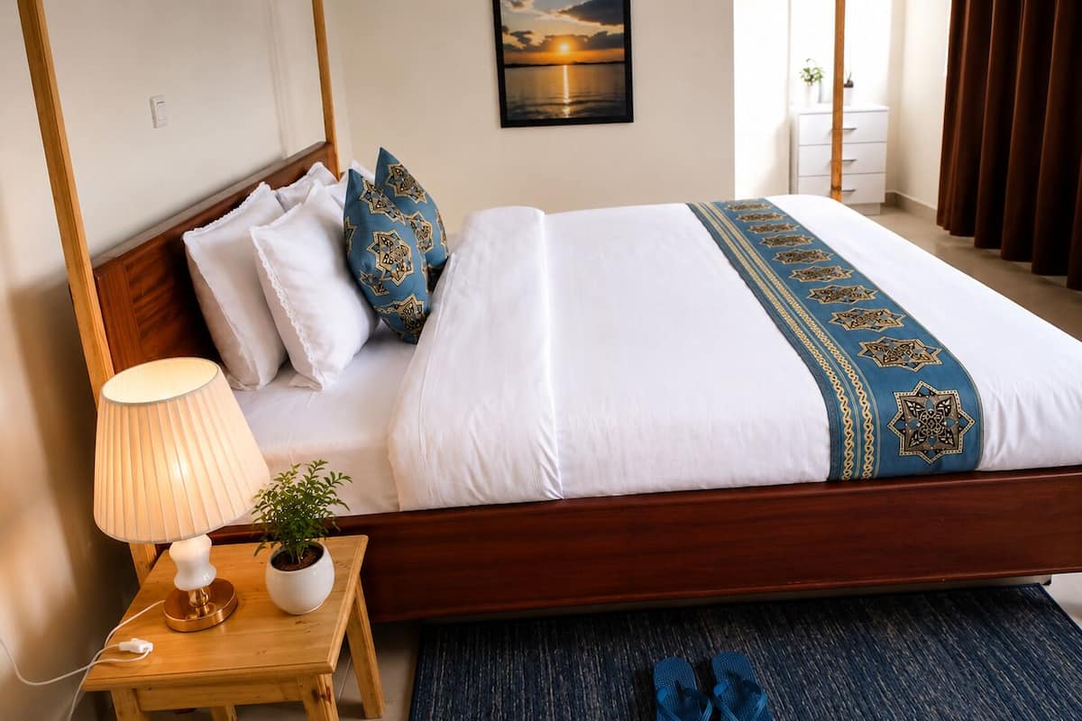 Una habitación de hotel con una cama de madera, ropa de cama blanca, almohadas decorativas, una lámpara de noche, una mesita con una planta y un par de pantuflas en el suelo.