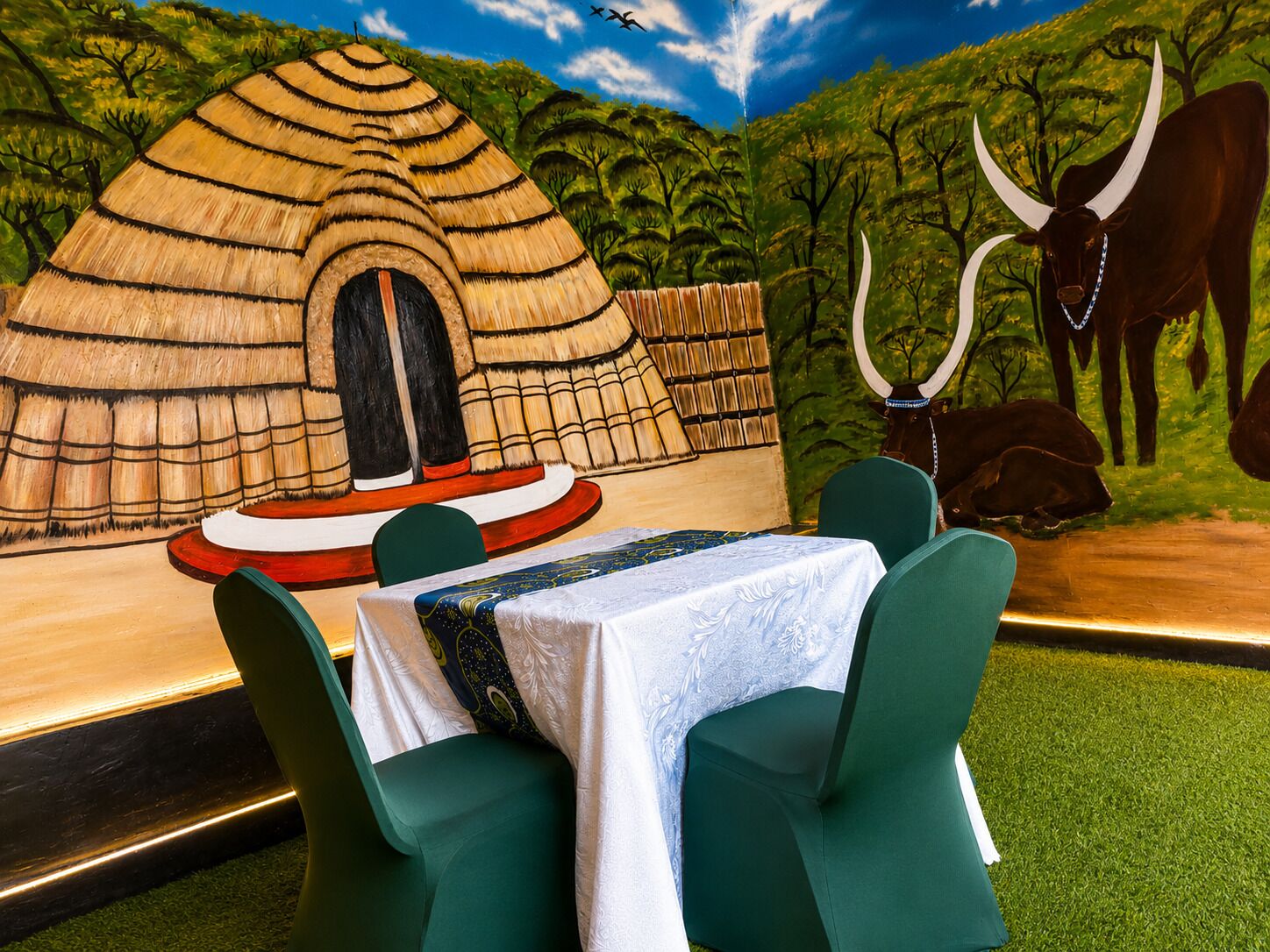 A dining area with a mural of a traditional hut, a table with a white tablecloth, and green chairs.