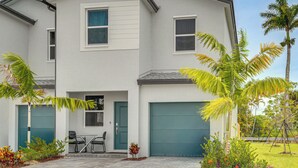 Une maison à deux étages avec un garage, une petite terrasse avec des chaises et une pelouse agrémentée de palmiers.