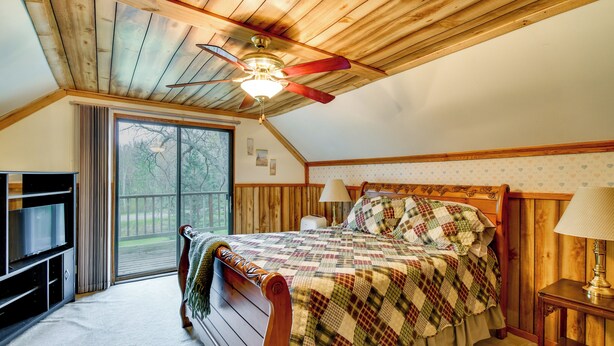 A bedroom with a wooden ceiling, a bed with a quilt, a TV, a lamp, and a view of the outdoors. - Classic Up North Cabin: Boat, Fish, ATV & Explore (Harrison)
