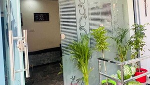 Entrada de edificio con puerta de vidrio, una zona interior con plantas en macetas y una escalera que conduce a la entrada.