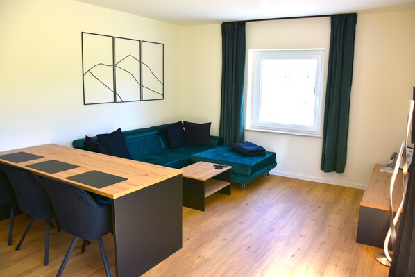 Un salon moderne avec une table à manger en bois, des chaises noires, un canapé de couleur turquoise et une grande fenêtre avec des rideaux.