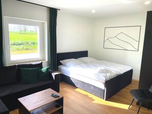 Un dormitorio con cama, sofá, una mesita, una silla, una ventana con vista a la vegetación y una obra de arte abstracto colgada en la pared.